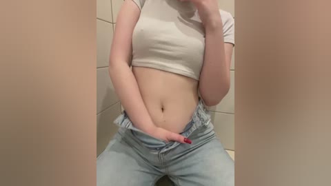 Video of a light-skinned woman with a slim figure, wearing a tight white crop top and light blue jeans, sitting in a tiled bathroom, with her hand on her crotch.