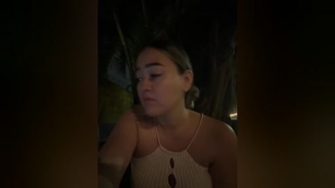 Video of a young woman with light skin, wearing a beige, ribbed tank top, slightly blurred in the foreground. Background shows a dimly lit, tropical outdoor setting with greenery and a soft, warm light.