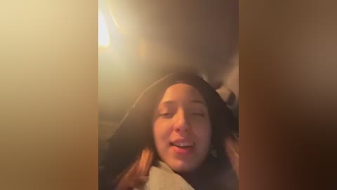 A video of a young woman with light brown hair, wearing a dark hoodie, lying on a bed, smiling with closed eyes, warm lighting creating a cozy ambiance.