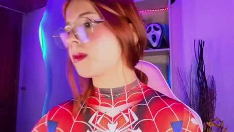 Video of a fair-skinned, red-haired woman in a Spider-Man suit, with glasses, red lipstick, and a neutral expression. Background features a white wall, bookshelf, and Halloween decorations.