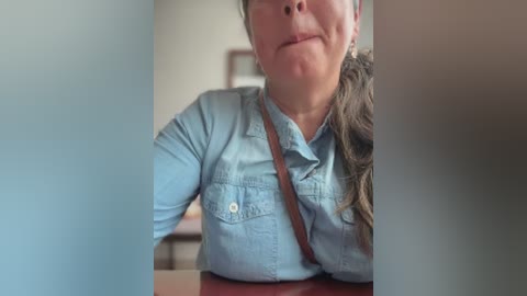 Media: Video of a woman with light skin, brown hair, and glasses, wearing a light blue denim shirt with a brown strap, smiling slightly.