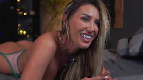 Media: Video of a smiling, topless blonde woman with medium-sized breasts, wearing a green thong, lying on a bed, surrounded by dimly lit, cozy indoor setting.