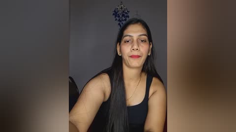 Video of a young woman with long black hair, wearing a black tank top, standing indoors with a dark background and a decorative blue ornament hanging above.