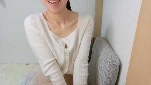 Video of a smiling Asian woman with light skin, wearing a white cardigan over a pink top, leaning against a gray fabric chair in a minimalist room with light walls.