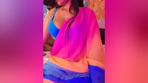 Video of a South Asian woman with long black hair wearing a vibrant blue bra and a pink-orange saree, posing indoors against a pink and purple gradient background.