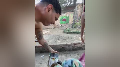 Video of a shirtless, tattooed man with short dark hair, crouched, sorting through scattered magazines on a dirt ground. Behind him, a partially visible green wall and brick building with a pink door.