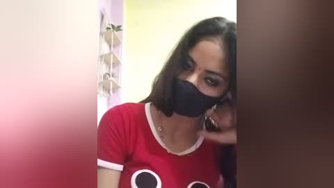 Video of a young woman with long dark hair, wearing a red t-shirt and black face mask, peeking through a doorway.