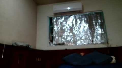 Media: A dimly lit bedroom with a large window covered in silver foil, an air conditioner mounted on the wall, and a dark wooden dresser below.