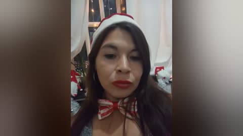 Video of a woman with long dark hair, wearing a Santa hat, red lipstick, and a red bow tie, indoors with festive decor.