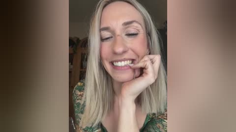 Video of a smiling, fair-skinned woman with blonde hair, wearing a floral blouse, touching her chin, in a warmly lit room with bookshelves in the background.