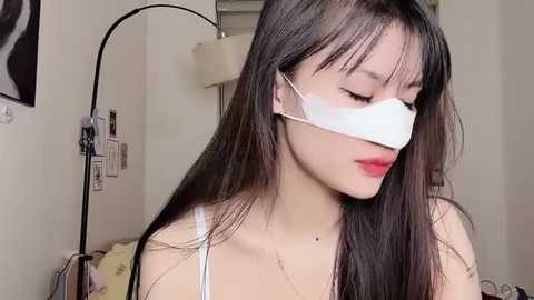 Video of a young Asian woman with long black hair, wearing a white surgical mask and a light pink top, in a simple, sparsely decorated room.
