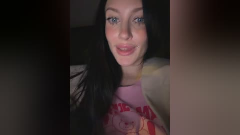 Video of a smiling woman with long black hair, wearing a pink tank top with a cartoon character design. The background is dimly lit, suggesting an indoor setting.