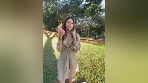 Video of an Asian woman in a beige trench coat, standing on a sunny, grassy park path with trees in the background. She has long dark hair and is smiling.