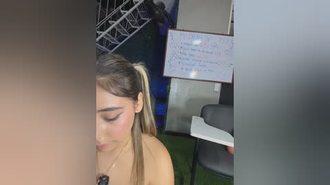 Video of a young Latina woman with long, straight, light brown hair in a ponytail, wearing a tan top. She's seated at a table in a classroom with a whiteboard, chairs, and green carpet.