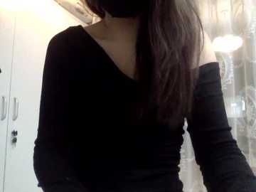 A video of a woman with long dark hair wearing a black off-shoulder top, partially obscured by a face mask, in a brightly lit room with white cabinets and a glass door in the background.