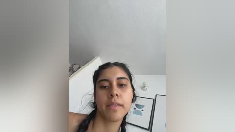 A video of a young South Asian woman with medium skin tone, dark hair, and a neutral expression, standing in a brightly lit room with white walls and minimalistic decor.