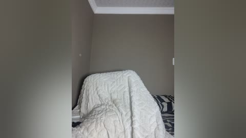 A video of a small, dimly-lit bedroom corner with a beige wall, white quilted blanket, black-and-white patterned pillow, and a white ceiling.