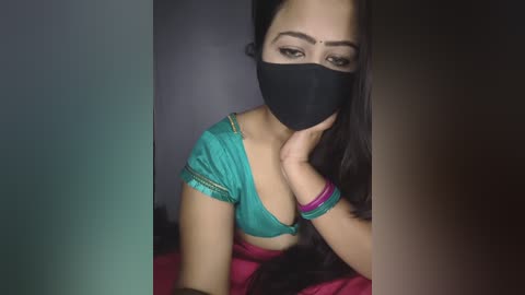 Video of a South Asian woman with long black hair, wearing a black face mask, turquoise blouse, and pink skirt. She's indoors with muted lighting, her expression serious and eyes partially hidden.