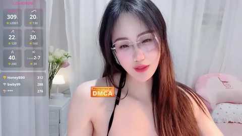 A video of an East Asian woman with long black hair, wearing glasses and a black top, in a soft-lit bedroom with pink bedding and white curtains.