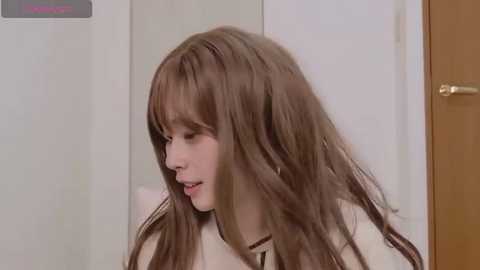 Video of a young Asian woman with long, straight brown hair, fair skin, and a light brown sweater, standing indoors against a plain white wall.