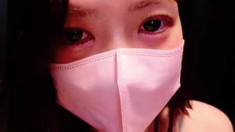 Close-up video of an East Asian woman with light skin, wearing a pink surgical mask that covers her nose and mouth, revealing her dark eyes and a hint of her dark hair.