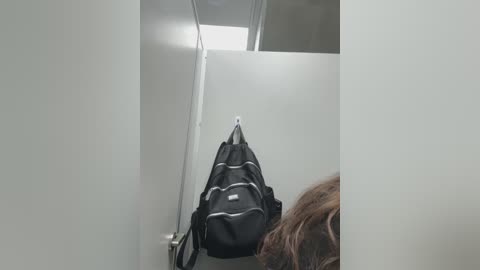 Video of a narrow, white-walled restroom stall with a black, striped gym bag hanging on the wall. A person with wavy brown hair is visible on the right, partially obscured by the bag.