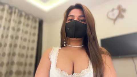 Video of a woman with long brown hair, wearing a black mask, white lace bra, and pearl necklace, standing in a bedroom with patterned curtains and a flat-screen TV.