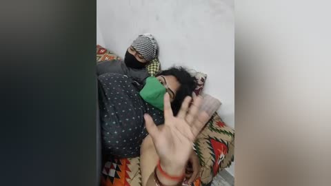 Media: Video of a woman with black hair, wearing a green mask and patterned gray dress, lying on a bed with a beige and green blanket, surrounded by a white wall.