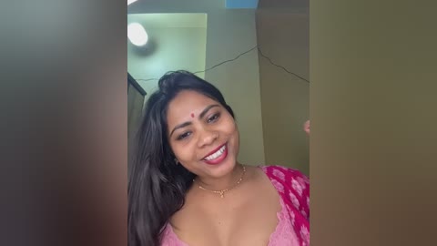 Video of a smiling South Asian woman with long black hair, wearing a pink floral blouse and a gold necklace, standing in a narrow corridor with beige walls.