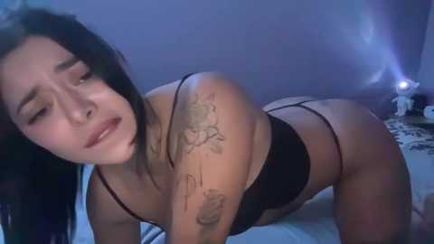 Video of a young woman with long black hair, wearing black lingerie, positioned on all fours on a bed, against a blue wall. She has a distressed expression, with visible tattoos on her arm and back.