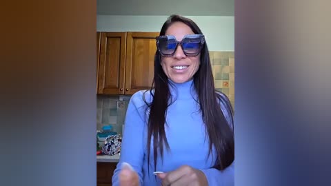 Video of a smiling woman with long, straight dark hair, wearing blue sunglasses and a light blue turtleneck sweater, in a kitchen with wooden cabinets and tiled backsplash.