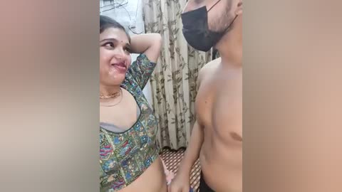 Video of a smiling South Asian woman with dark hair, wearing a colorful crop top, and a black face mask, standing with a shirtless man, in a room with floral curtains.
