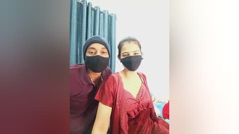 Video of two masked Asian people in a hospital room with blue curtains, a man in a dark shirt, and a woman in a red dress, both looking forward.