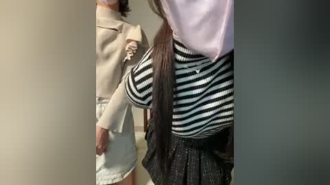 Video of two children, one in beige long-sleeve top, the other in black-and-white striped top and grey skirt, standing side by side.