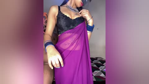 Video of a light-skinned woman with dark hair, wearing a sheer purple sari over a black lace bra, holding the sari with blue bracelets. Background shows a patchwork quilt.