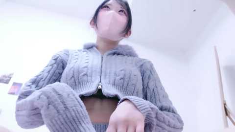 A video of an Asian woman with dark hair and fair skin wearing a grey, knitted cropped sweater, looking upward. Background includes white walls and a blurred painting.