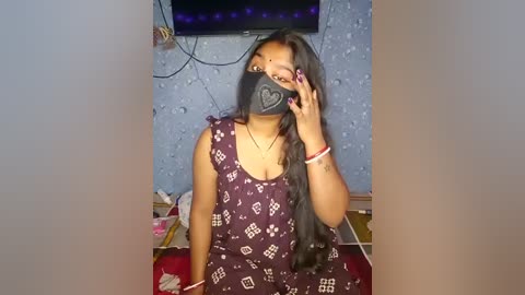 Video of a young South Asian woman with long black hair, wearing a purple patterned dress, black mask with a heart design, and red bracelets, standing indoors with blue textured wallpaper, a TV, and cluttered shelves in the background.