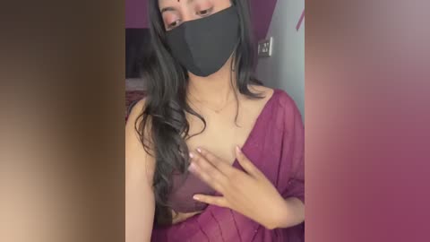 Video of a South Asian woman with long black hair, wearing a black face mask, a deep red off-shoulder dress, and a black top, covering her breasts with her left hand. Background shows a blurred pink wall and a doorway.
