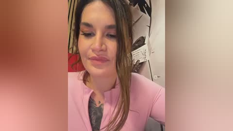 Media: A video of a young woman with medium skin tone, long brown hair, wearing a pink V-neck top, and subtle makeup. The background features a cluttered room with a white door and hanging clothes.