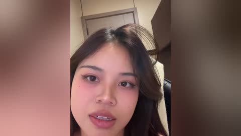 Video of a young Asian woman with fair skin, long black hair, and dark eyes, smiling slightly, wearing a beige top, in a beige room with a door in the background.
