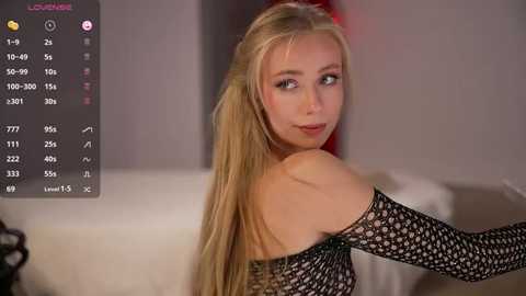 Video of a blonde Caucasian woman with fair skin, wearing a black fishnet top, sitting on a white couch in a dimly-lit room.