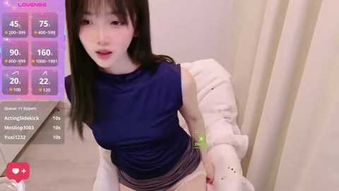 Video of a young Asian woman with long black hair, wearing a sleeveless dark blue top and white shorts, in a modern indoor setting.