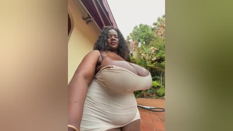 A video of a plus-size Black woman with large breasts wearing a tight beige tank top and shorts, standing outside near a house with a garden.
