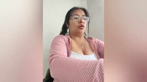Video of a middle-aged Asian woman with long black hair, wearing glasses and a pink textured cardigan over a white top, seated indoors against a plain white wall.