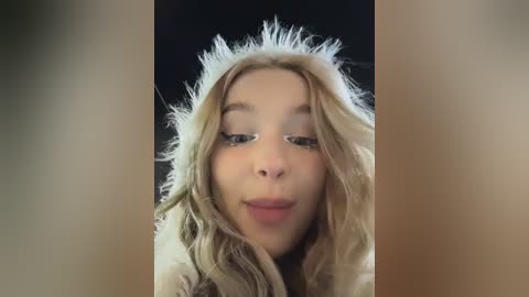 A close-up video of a young woman with long, wavy blonde hair, wearing a white, fluffy headband, against a black background. Her face is partially obscured by her hair.