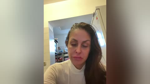 Video of a woman with long, dark brown hair in a white long-sleeve shirt, looking downward, in a bathroom with white tiles and shelves, blurred in the background.