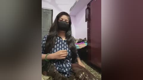 Media: Video of a woman in a mask, wearing a blue patterned shirt, seated on a bed with a maroon headboard. The room is cluttered with clothes, and the walls are pale pink.