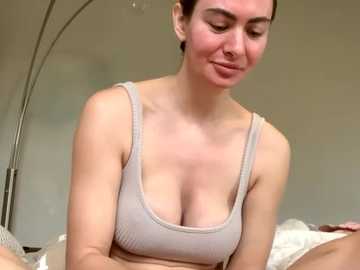 Video of a light-skinned woman with brown hair in a beige sports bra, sitting on a bed with white sheets. She has a subtle smile. Background includes a metal floor lamp.