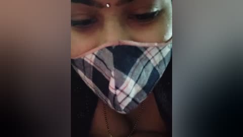 A close-up video of a woman's face, partially obscured by a blue and white plaid face mask. She has dark skin and wears a black jacket. The background is dimly lit, adding a moody atmosphere.