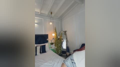 Video of a modern, minimalist bedroom with white walls, a cozy wooden ceiling, a small Christmas tree, and a bed adorned with white linens and navy blue pillows.